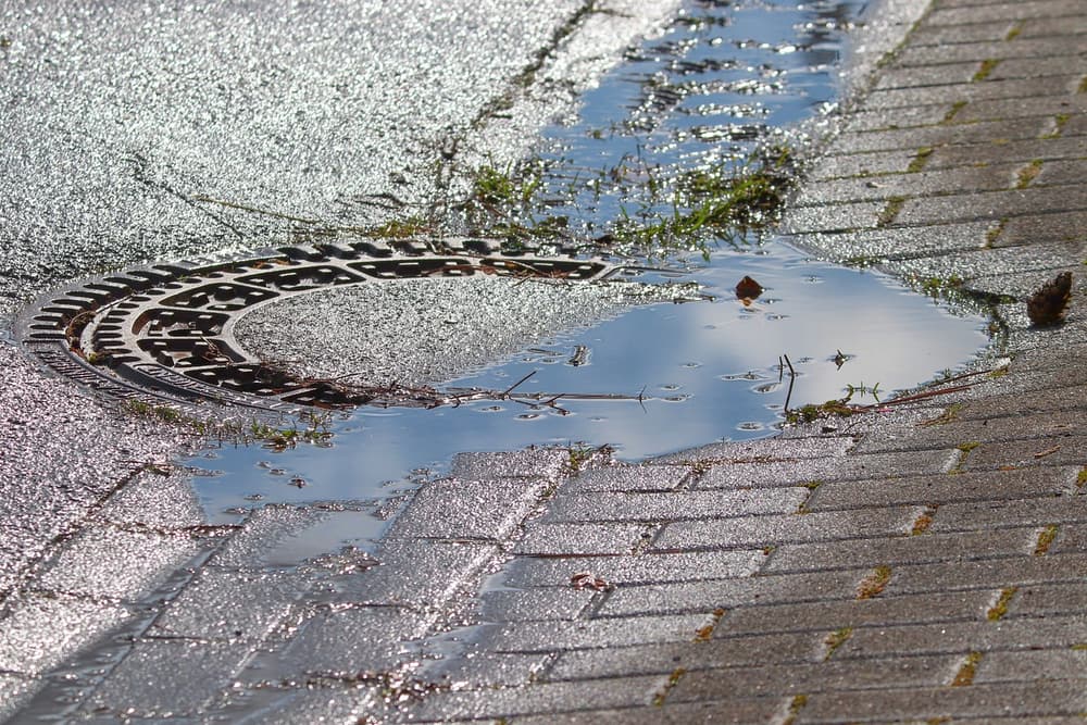 Runder gusseiserner Kanaldeckel im Pflaster, daneben eine Regenpfütze, in der sich der blaue Himmel spiegelt