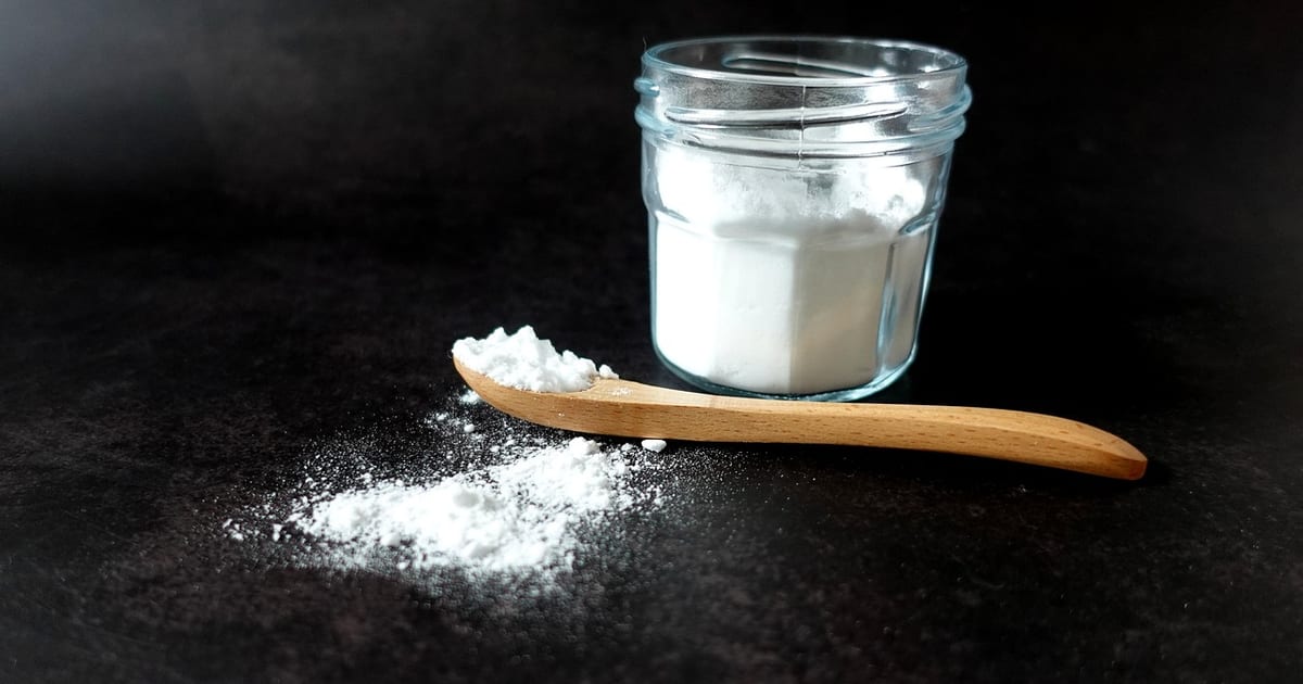 Weißes Natronpulver in einem Glas, davor ein Holzlöffel mit Pulver auf dunkler Arbeitsfläche