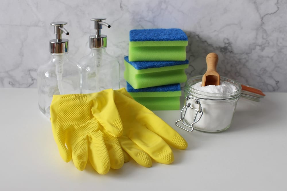 Gelbe Gummihandschuhe vor einem Stapel grün-blauer Schwämme, daneben ein kleines Bügelglas mit weißem Pulver und Holzlöffel und zwei Glasspender
