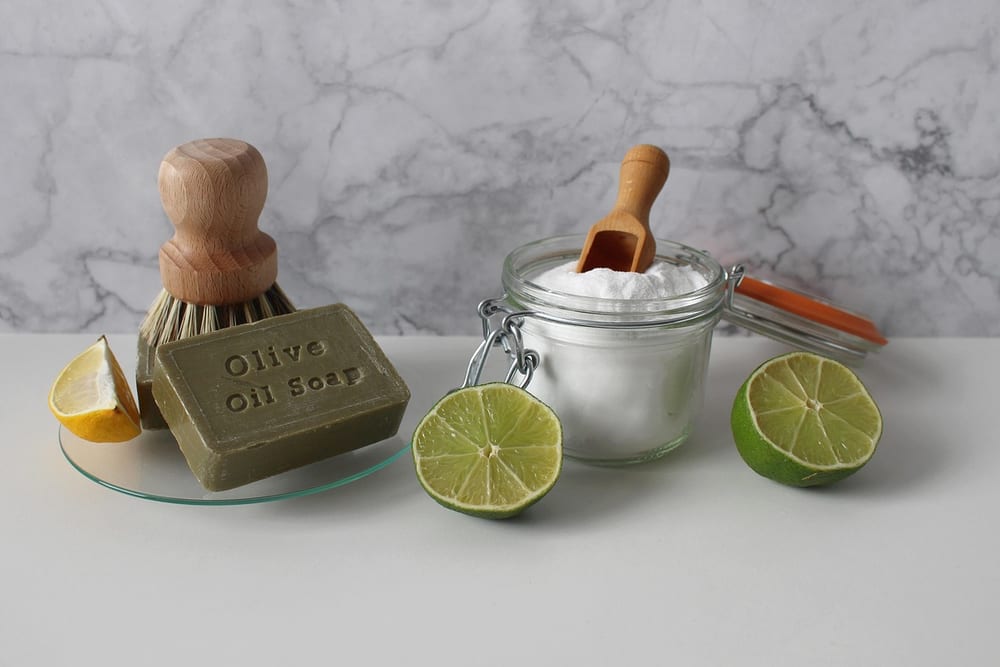 Bügelglas mit weißem Pulver und Holzlöffel neben einer Holzbürste, einem Stück Olivenölseife und mehreren Limettenhälften