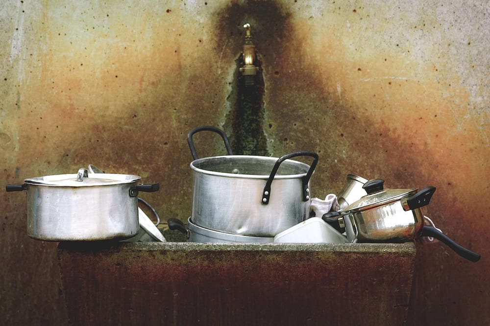Mehrere Kochtöpfe aus Edelstahl stehen auf einer Spüle vor einer gefliesten Wand mit Wasserhahn
